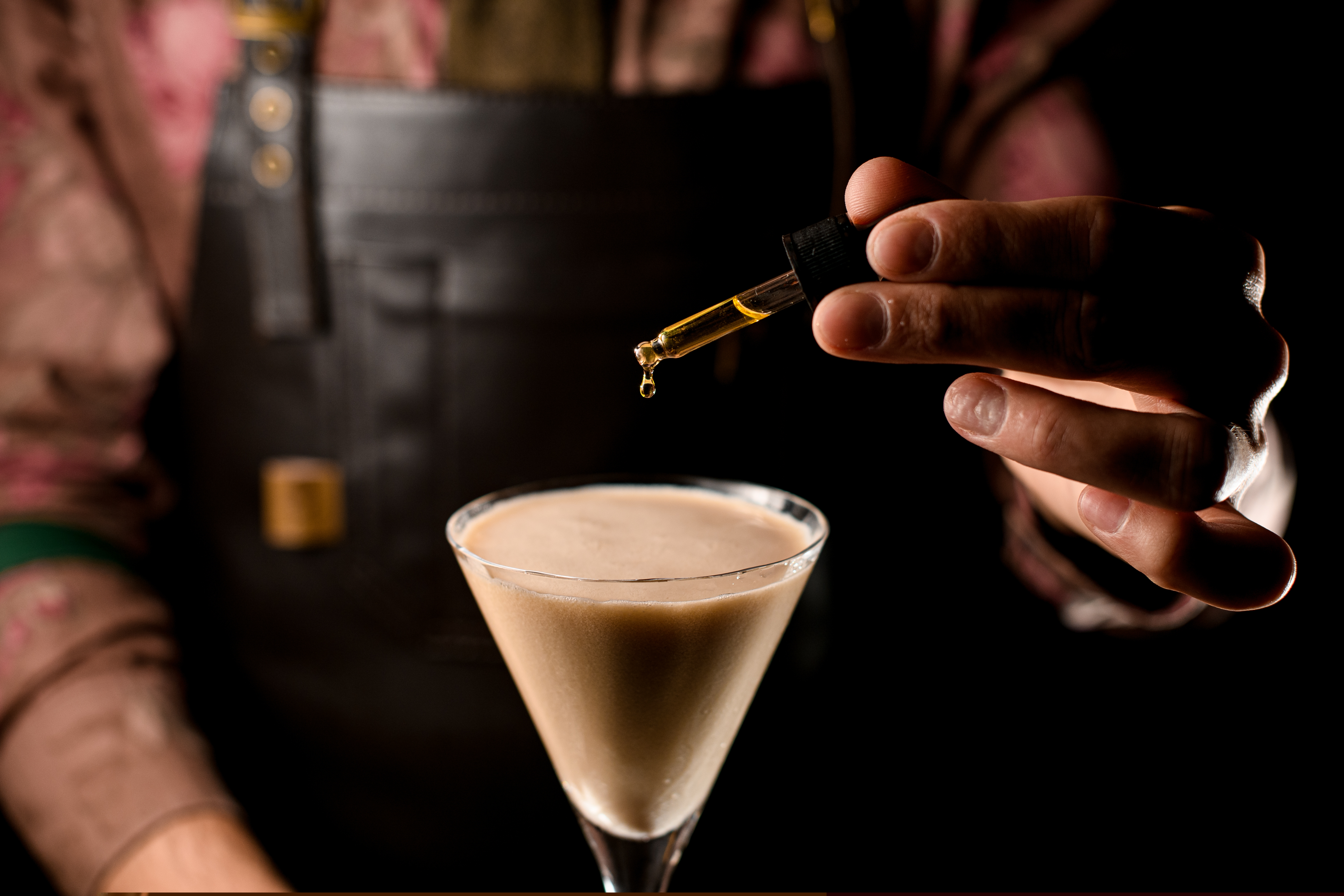 Man in a leather apron adding drops of bitters from a glass dropper into a cocktail in a martini glass