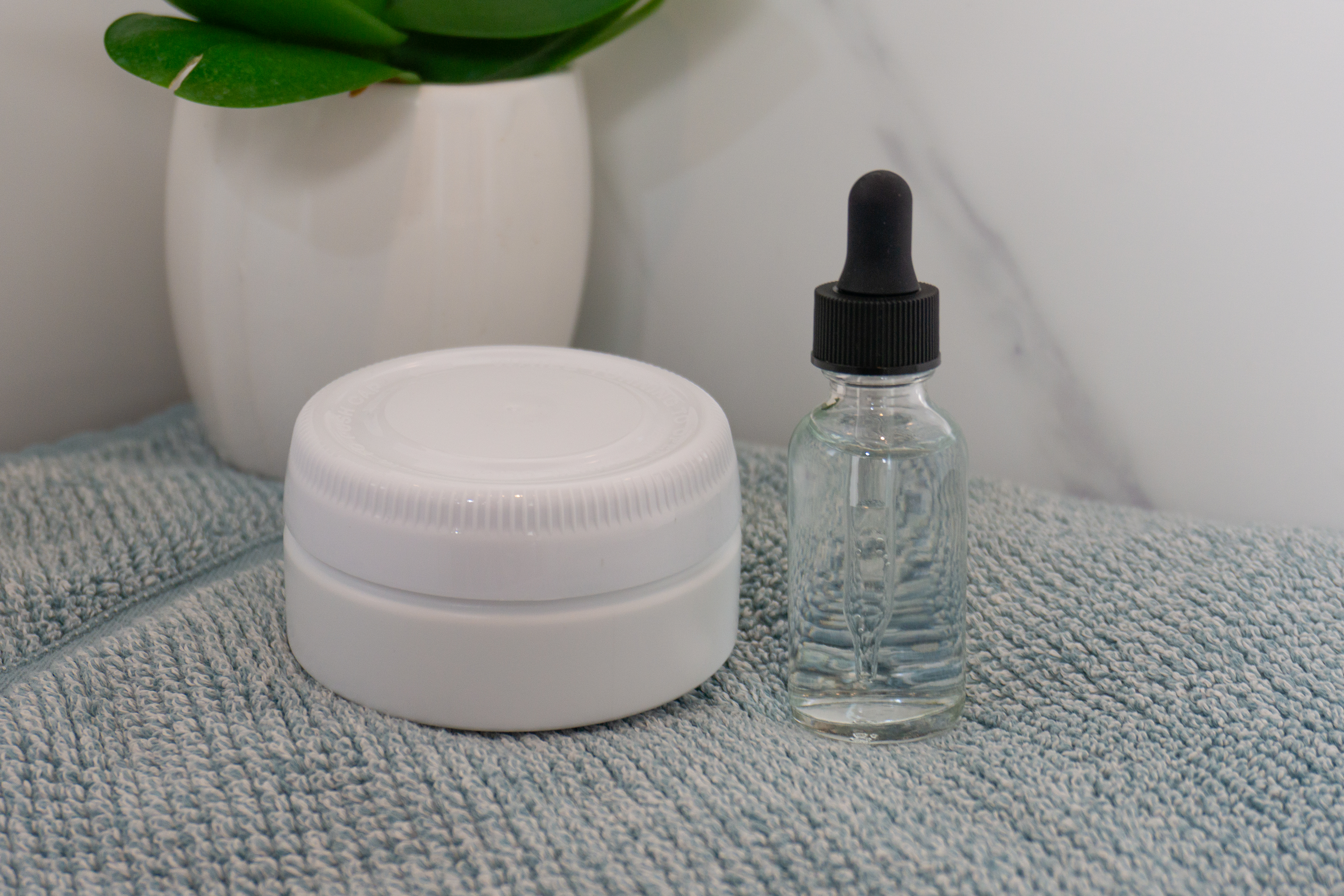 a boston round bottle with dropper cap on a blue towel next to a white tub of cream